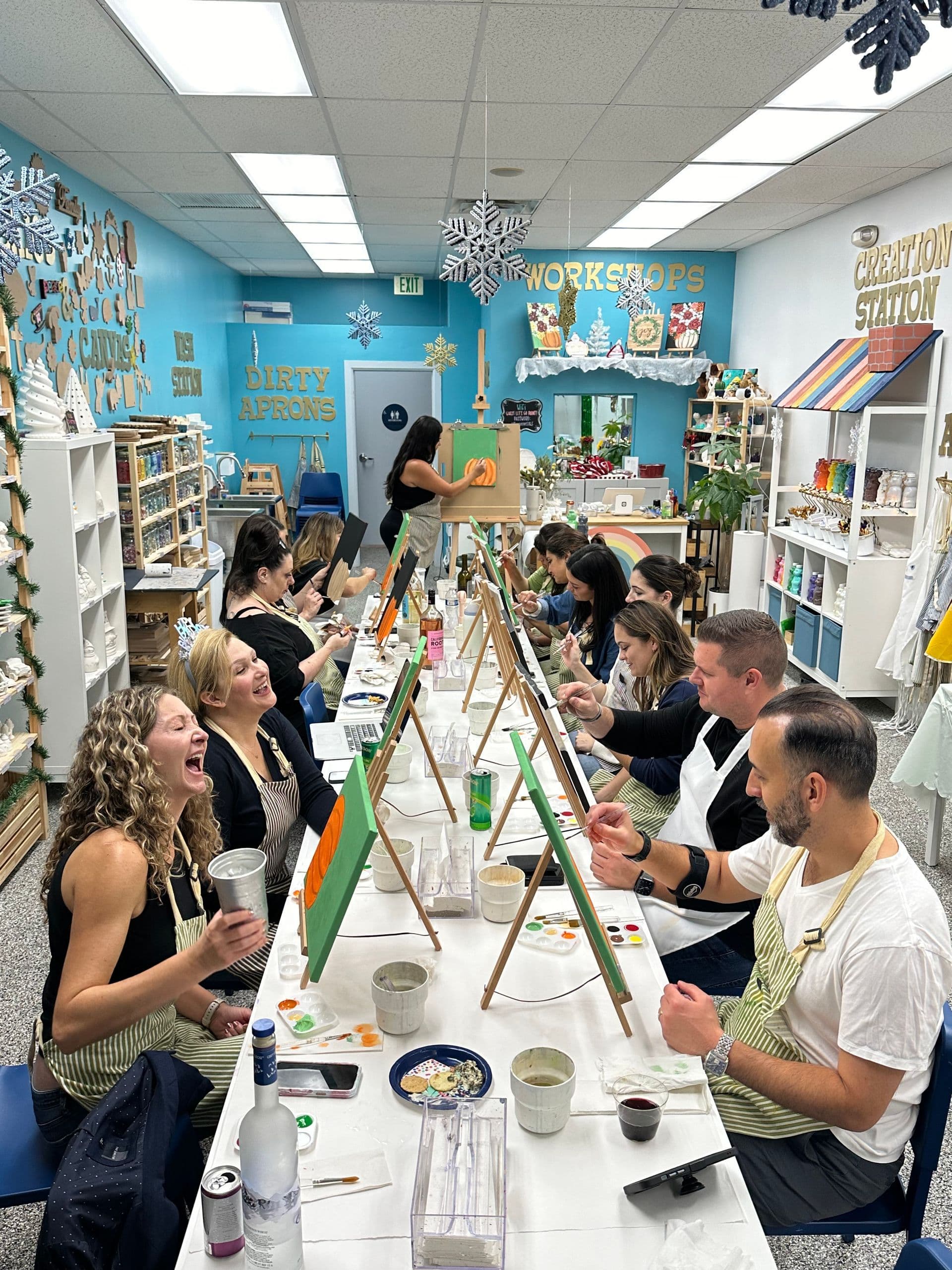 A long table of adults laughing through a paint-and-sip night at Let's Go Paint, easels in front of every guest.